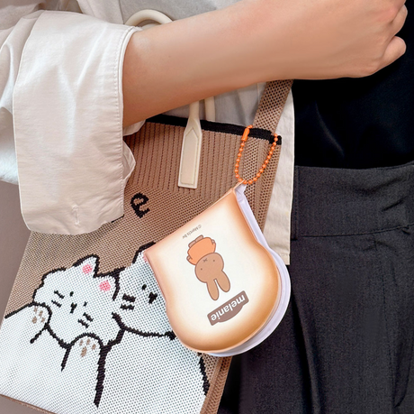 Hand holding tote bag with cat design and a toast-shaped memo pad keychain featuring a character named Melanie attached to the bag