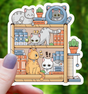 Hand holding a cute sticker of a bookshelf filled with playful cartoon cats and colorful books and plants