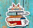 Sticker of an orange cat peeking over a stack of cozy books with a steaming cup and a small mouse on a blue wooden background