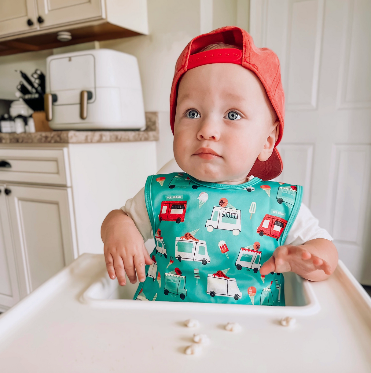 Baby wearing a colorful ice cream truck bib apron with full coverage sitting in a high chair in a kitchen