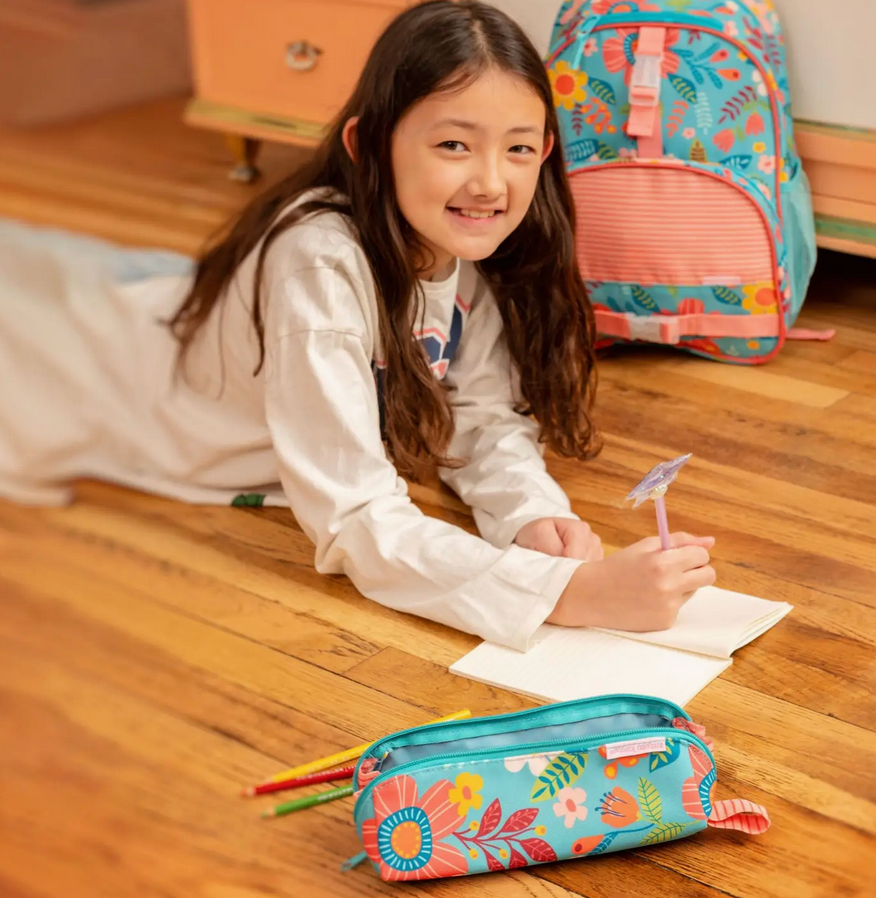 Smiling girl lying on wooden floor writing in notebook with colorful floral pencil pouch and matching backpack nearby