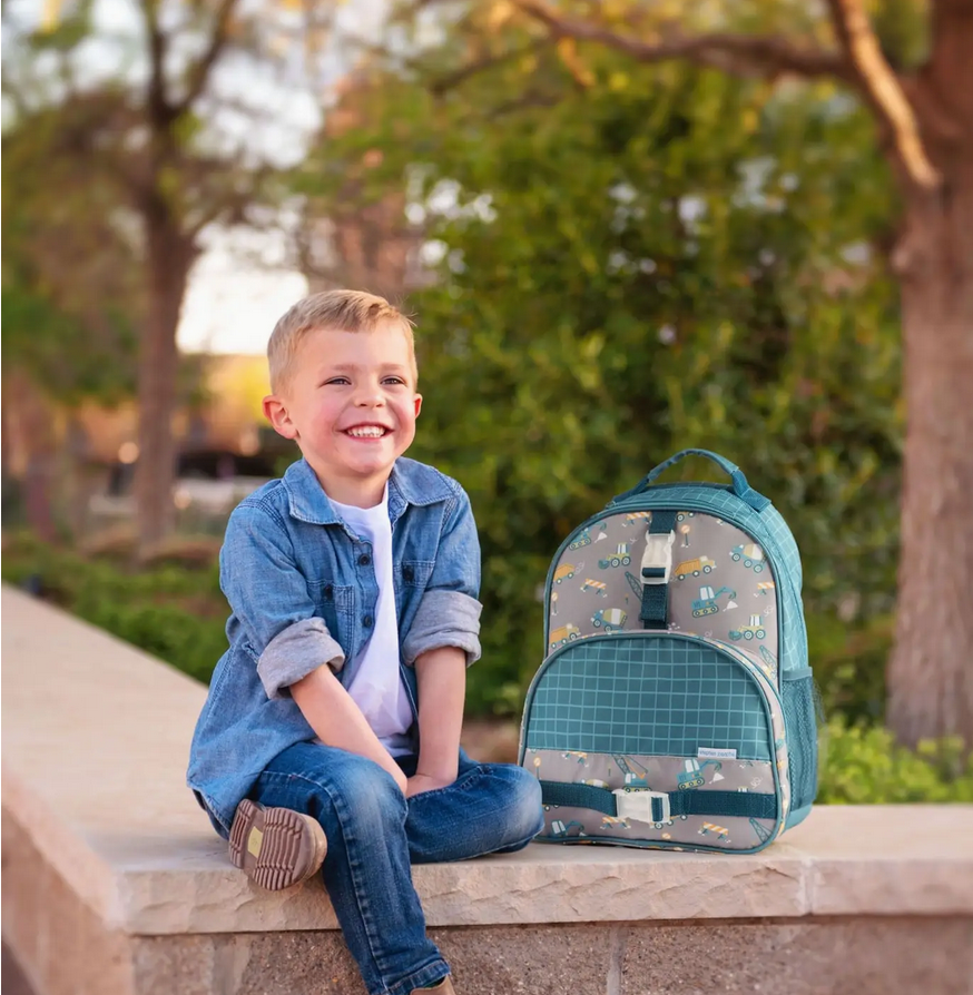 Smiling young boy sitting outdoors next to a gray and teal backpack with a bee and tractor print design
