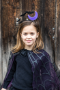Young girl wearing a Halloween headband with black bats flying around a purple crescent moon and a spider web cape