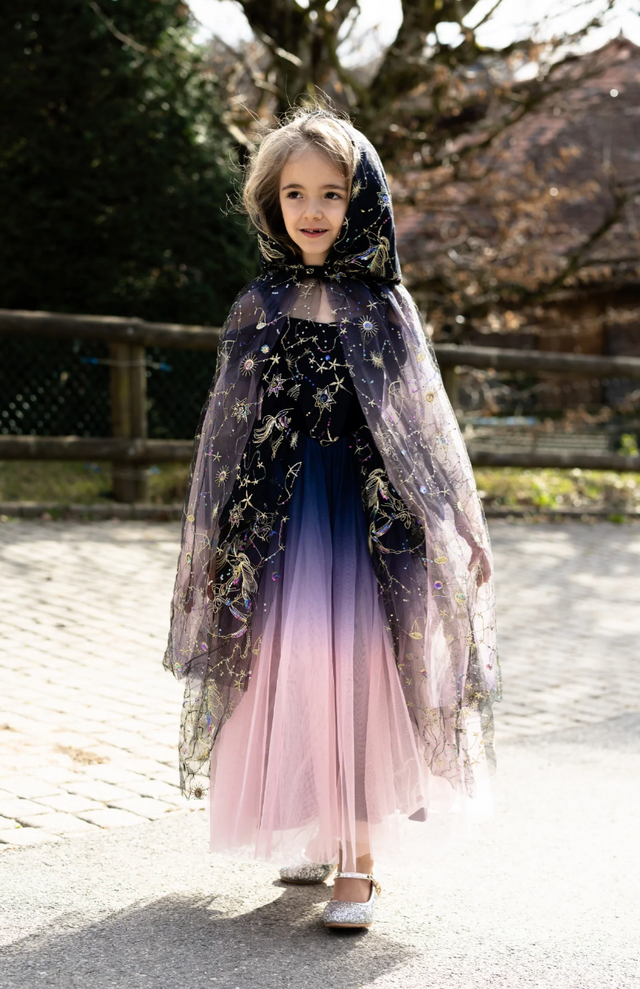 Little girl wearing a dark purple shimmering cloak with embroidered stars and a roomy hood outdoors on a sunny day.