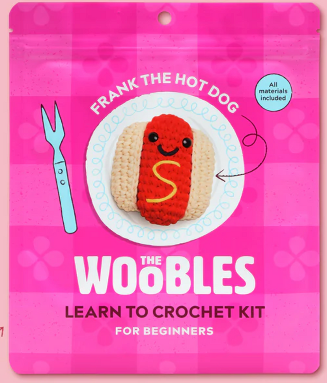 Bright pink packaging of The Woobles crochet kit featuring a smiling crocheted hot dog with mustard detail on a white plate design.