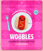 Bright pink packaging of The Woobles crochet kit featuring a smiling crocheted hot dog with mustard detail on a white plate design.