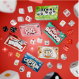 Mini Dice Game set with six colorful game boxes and various dice scattered on a red background for fun and travel play.