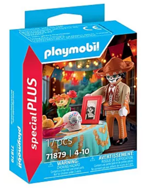 Playmobil figure with hat and Día de los Muertos table featuring skull, flowers, fruit, and framed photo for celebration.