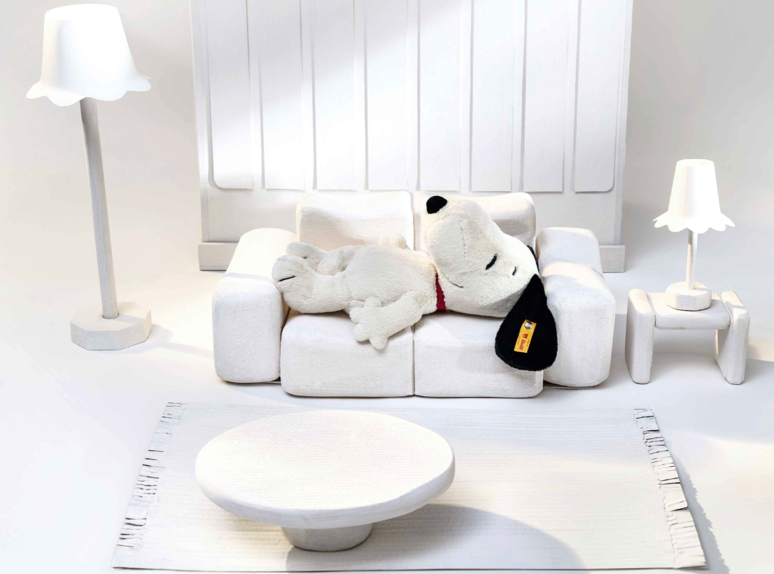 Soft plush Snoopy lying on a white couch in a minimalist all-white living room setting with lamps and table.