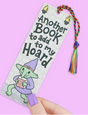 Bookmark with a cute goblin illustration holding a book and rainbow tassel on a light brick background