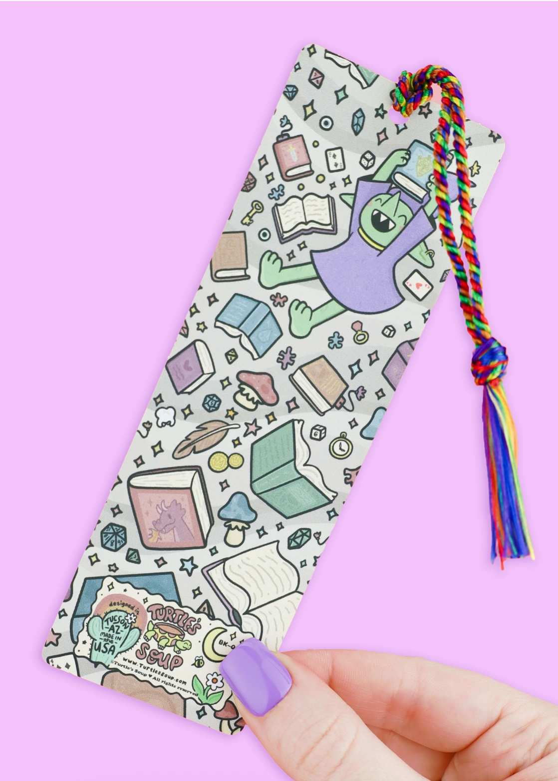 Illustrated bookmark with a joyful goblin, books, and colorful rainbow tassel on a purple background