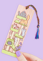 Bookmark featuring playful cats on a tower with a colorful rainbow tassel against a purple background