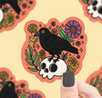 Hand holding a colorful sticker featuring a black crow perched on a white skull surrounded by bright flowers and leaves