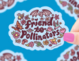 Hand holding colorful "friend to pollinators" sticker surrounded by bees, butterflies, flowers, and a bat on blue background