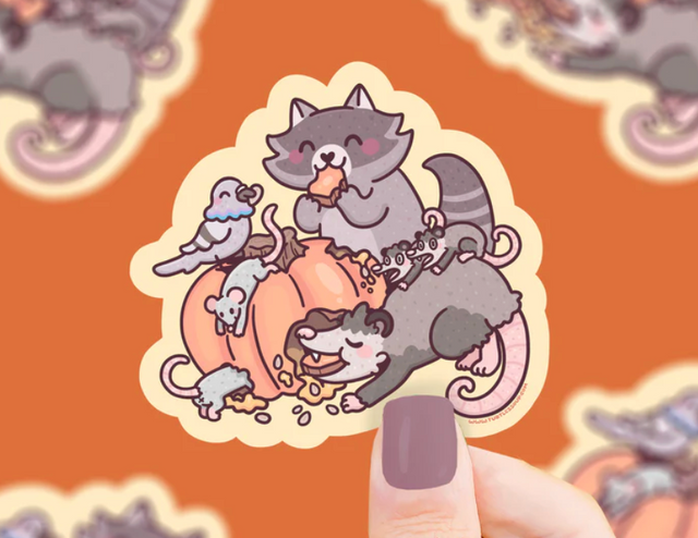 Hand holding a weatherproof sticker of trash animals happily feasting on a partially eaten pumpkin.