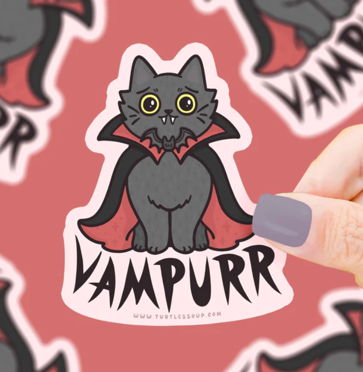 Hand holding a 3-inch vampire cat sticker with a red cape and big yellow eyes on a pink background.