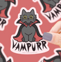 Hand holding a 3-inch vampire cat sticker with a red cape and big yellow eyes on a pink background.