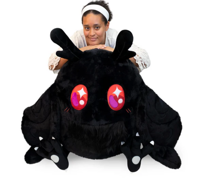 A person leaning on a large soft black Mothman plush with big colorful eyes and extended wings.