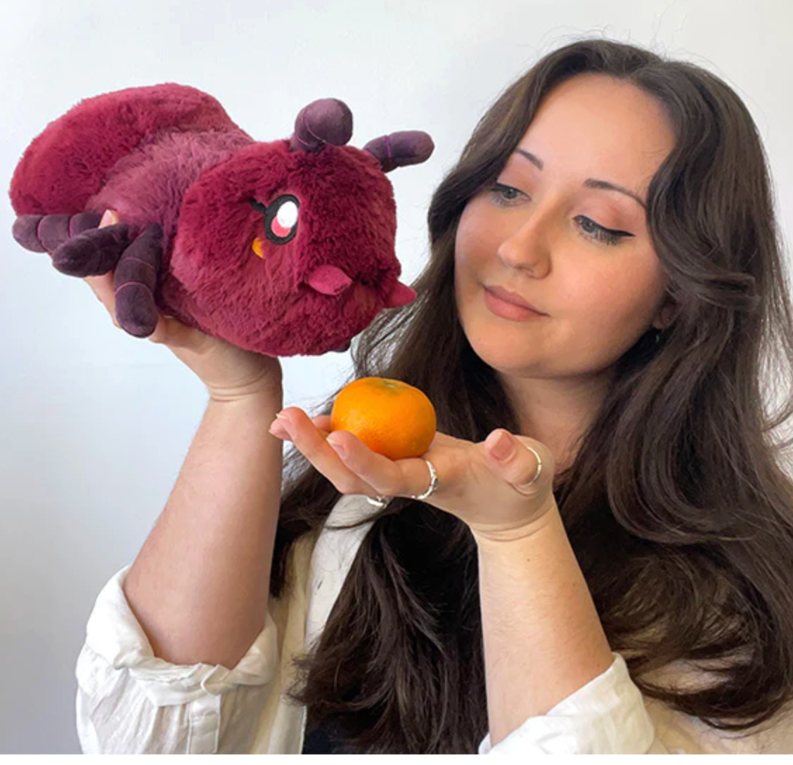 Woman holding a plush Ant Mini toy above her hand while looking at a small orange fruit with a gentle expression