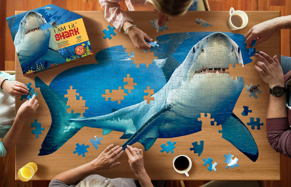 People assembling a 100-piece oversized jigsaw puzzle of a large friendly shark on a wooden table with beverage cups nearby