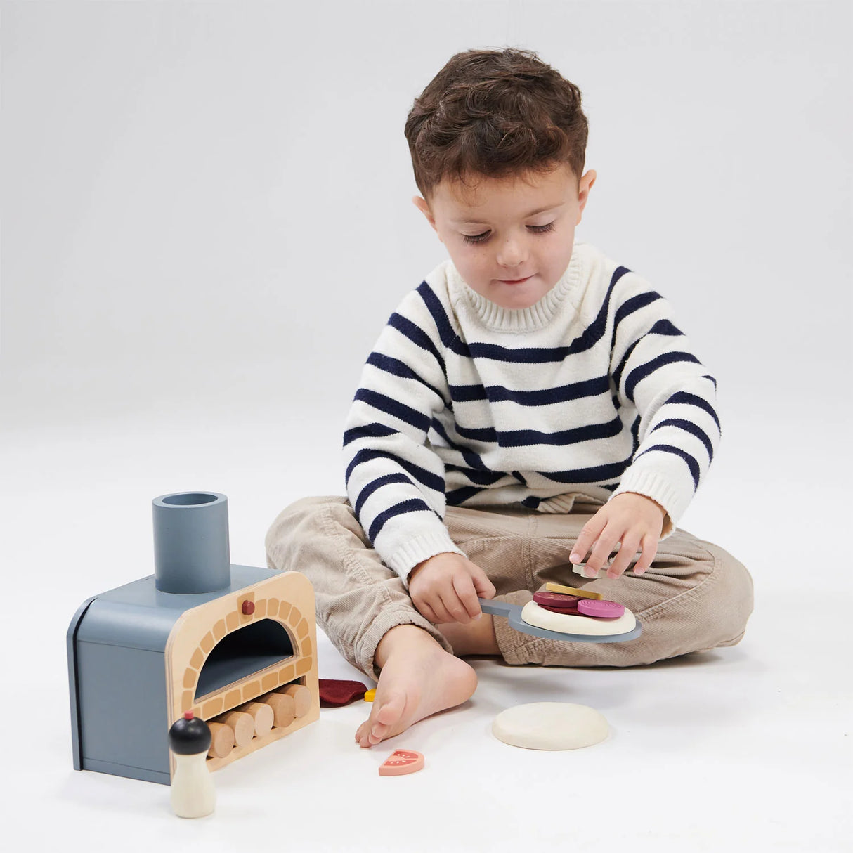 Child playing with pretend pizza oven and toppings using the Make Me a Pizza! toy set on a white background.