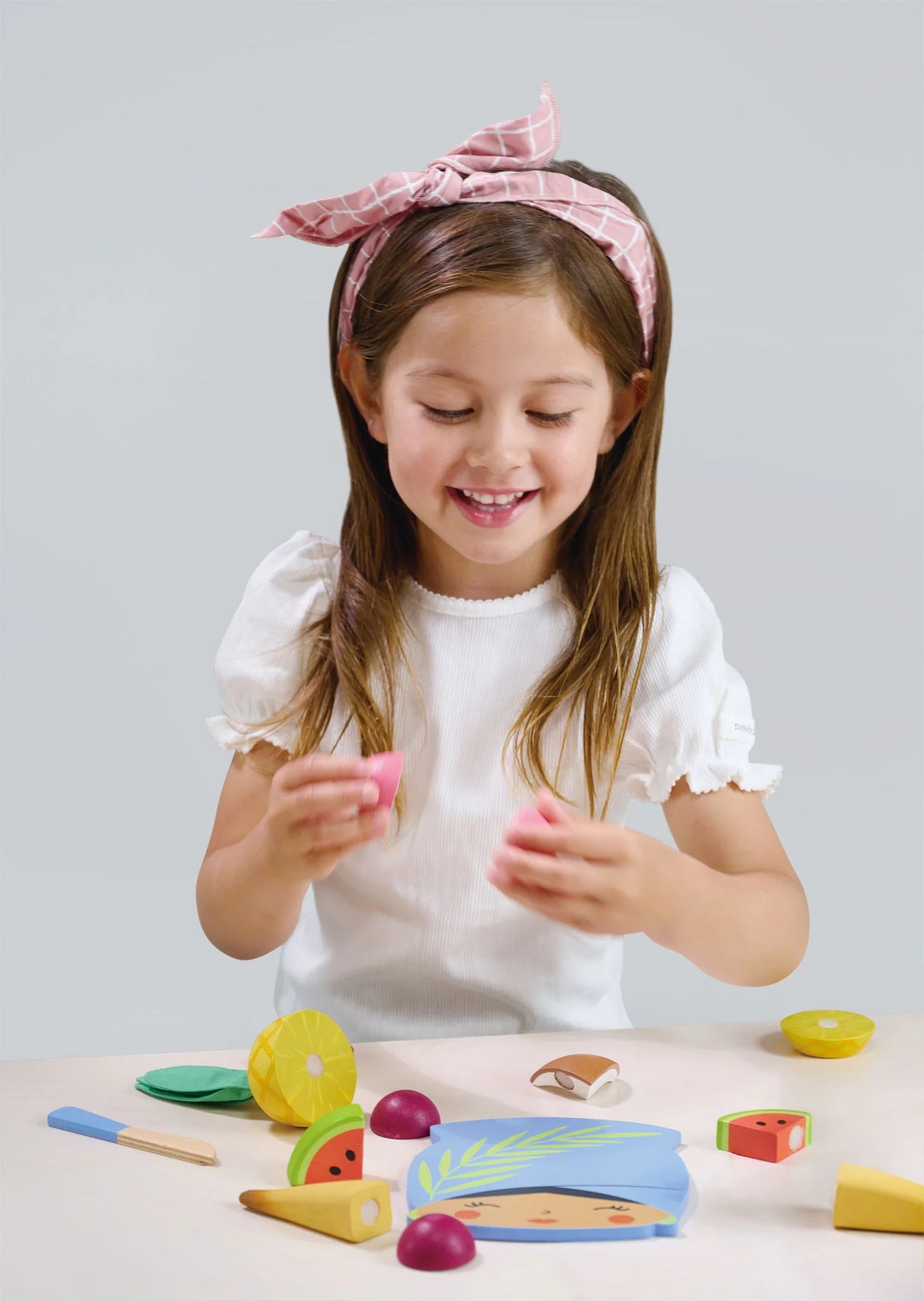 Child playing with Tropical Fruit Chopping Board wooden fruit pieces and blue knife on table.