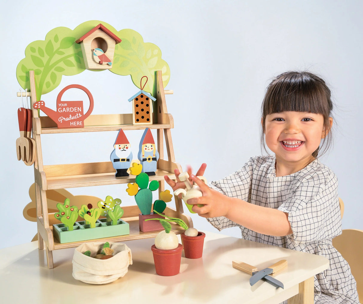 Garden Centre wooden playset with shelves, garden tools, pots, gnomes, and birdhouse, enjoyed by a smiling child.