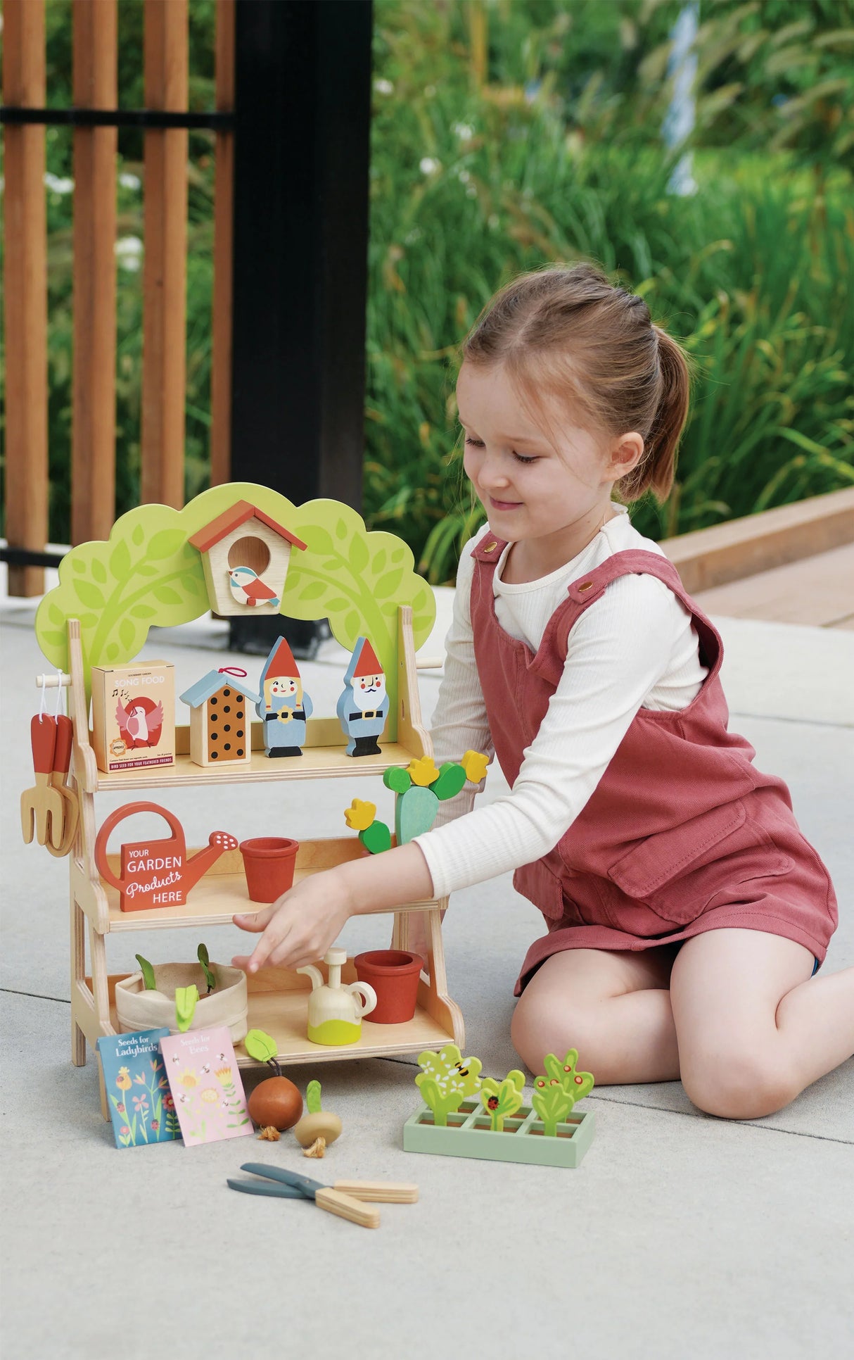 Young girl playing with the Garden Centre wooden stand featuring garden tools, birdhouse, gnomes, and plant accessories.