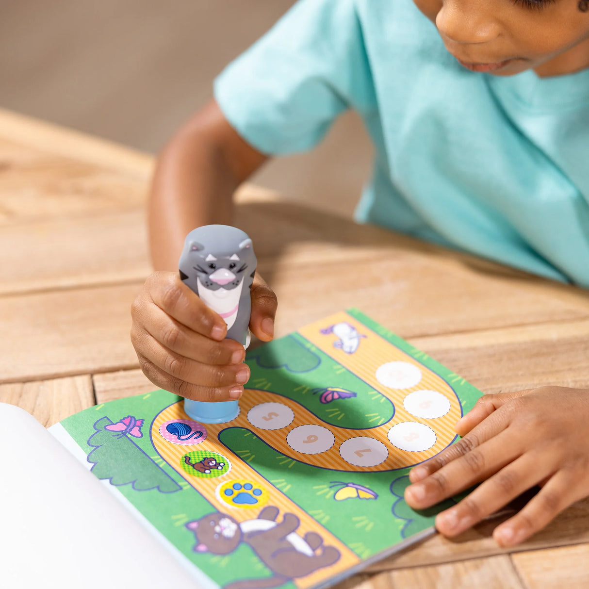 Child using the Sticker WOW! Cat to place stickers on a colorful activity book page at a wooden table.