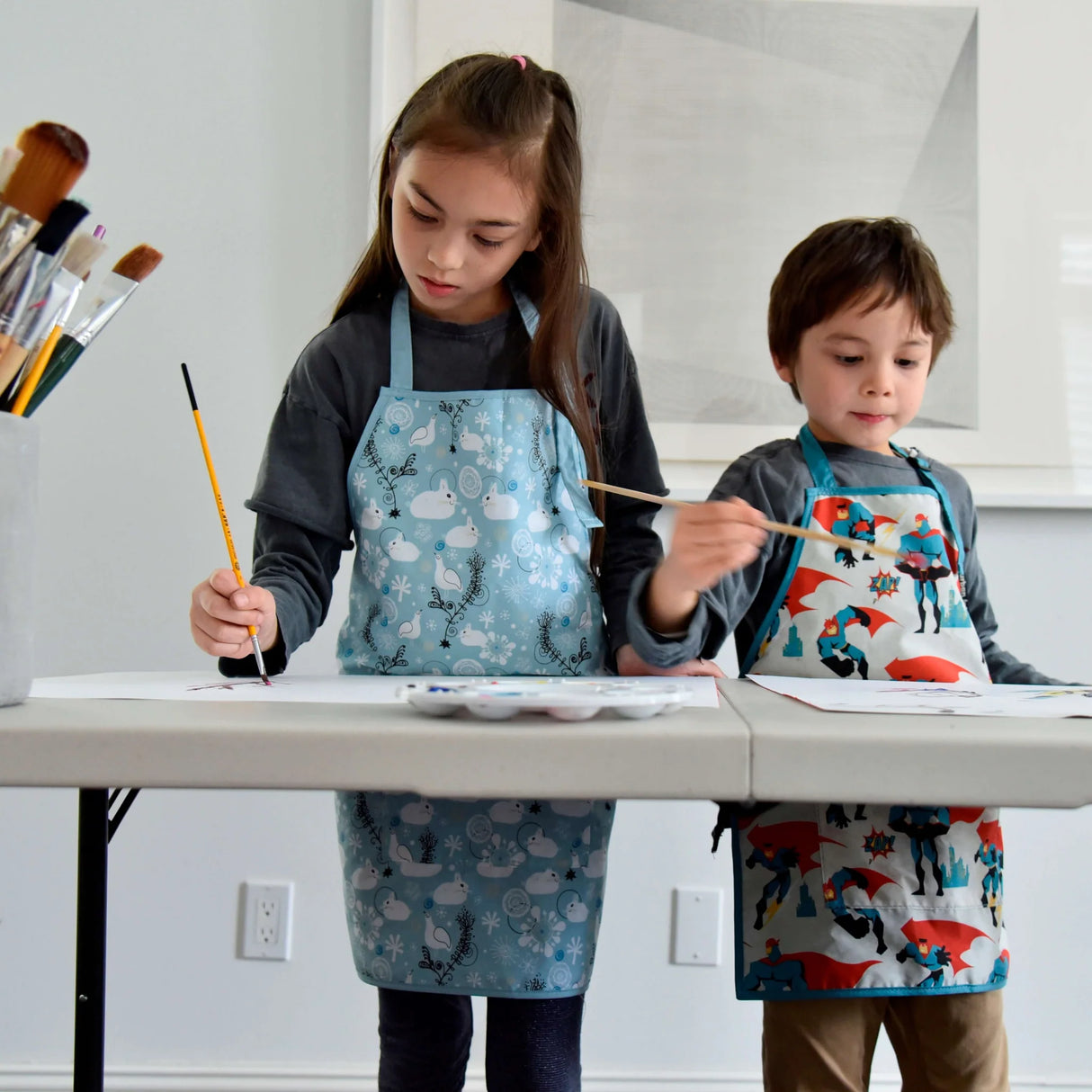 Children wearing Little Helper Apron | Bunnies Medium painting at a table, perfect for kids cooking and crafting.