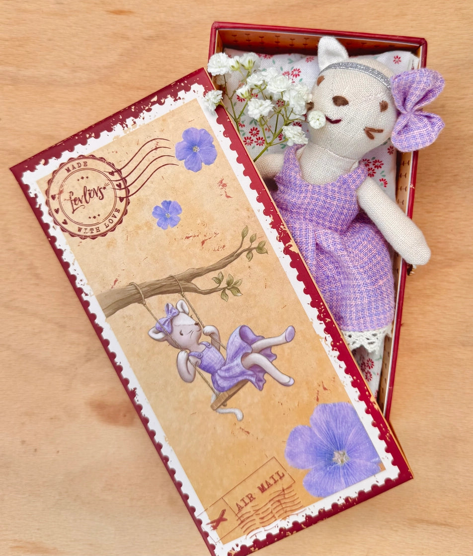 Cat doll in a purple dress with bow, resting inside a vintage floral stamp box with a cushioned interior.
