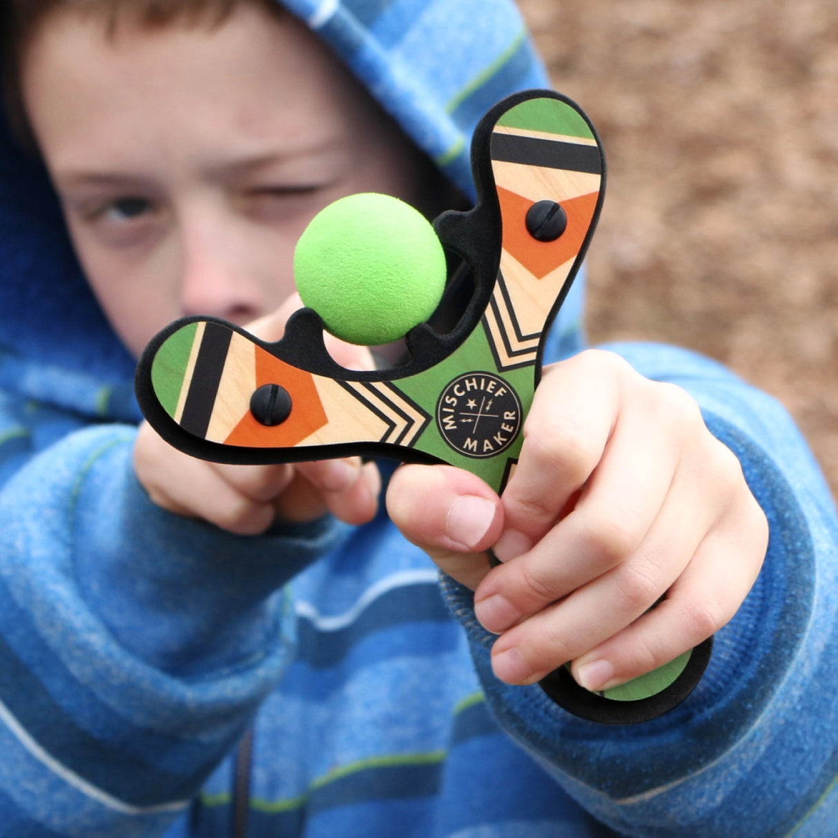 Child aiming a wooden Mischief Maker slingshot with a soft green foam ball ready to shoot outdoors