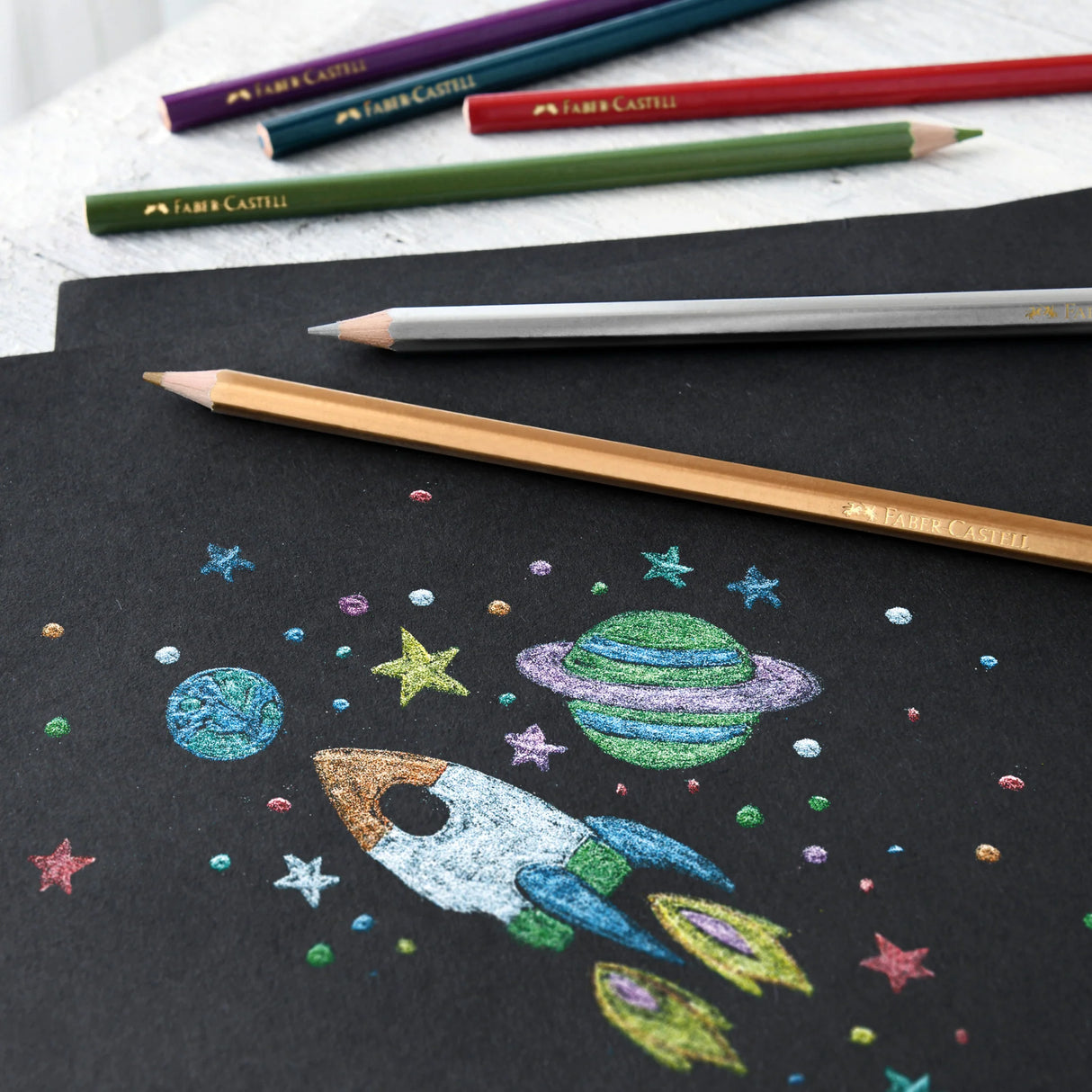 Metallic Colored Pencils with shimmering metallic colors drawing a vibrant rocket and planets on black paper.