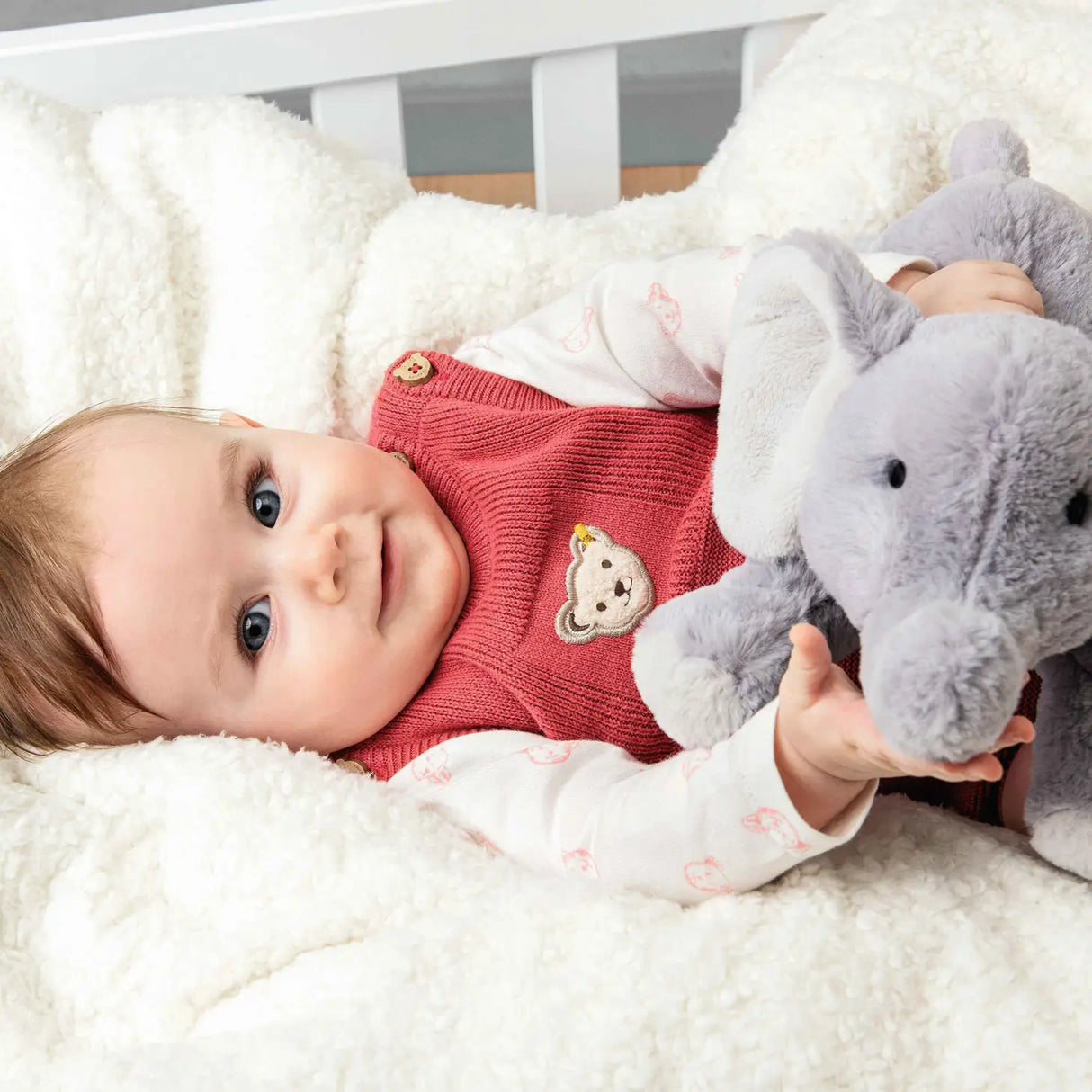 Baby lying on soft blanket holding Elephant Baby Elna plush toy with curved trunk and oversized ears.