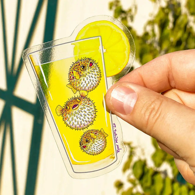Hand holding a transparent Spiked Lemonade Sticker featuring pufferfish floating in a lemonade glass with a lemon slice.