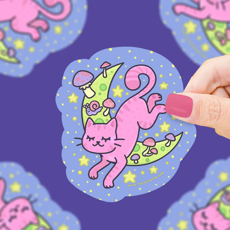 Hand holding a vibrant vinyl sticker of a pink cat jumping over a green crescent moon with stars and mushrooms.