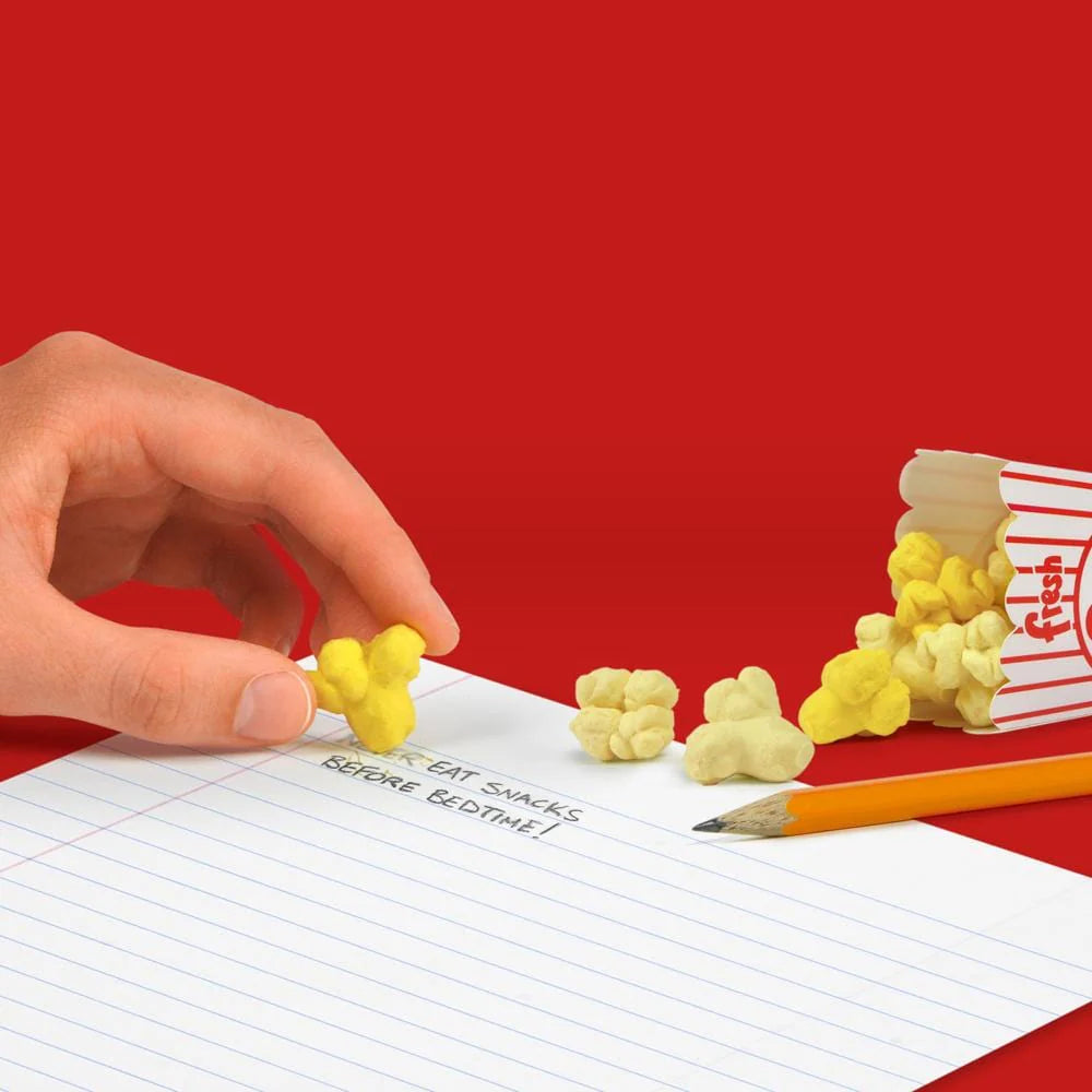 A hand holding a yellow eraser shaped like popcorn from a box of Fresh Popcorn Erasers on a notebook.