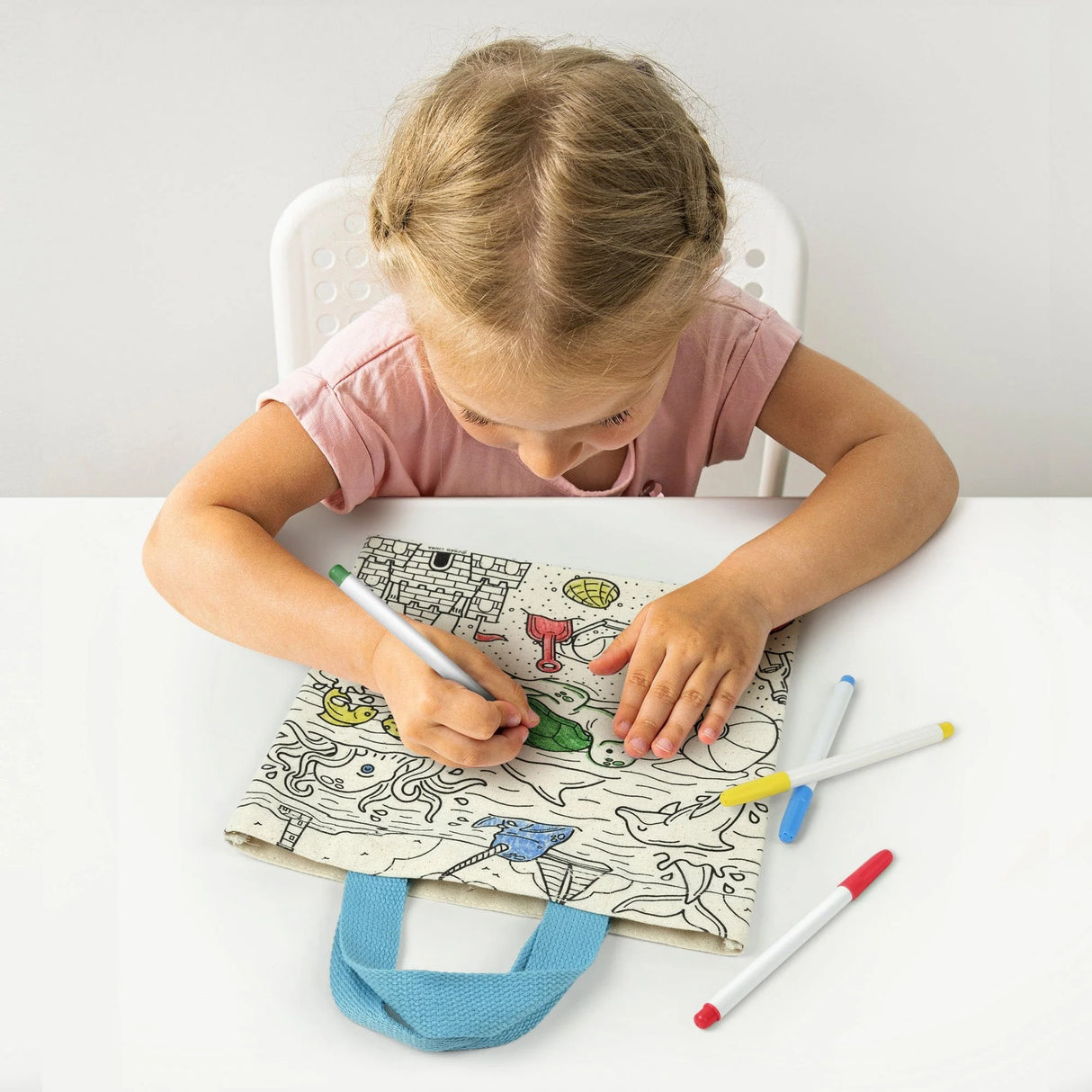 Young girl coloring a canvas tote bag with markers, showcasing the Kit & Cadoodle Color-In Tote Bag creative activity.