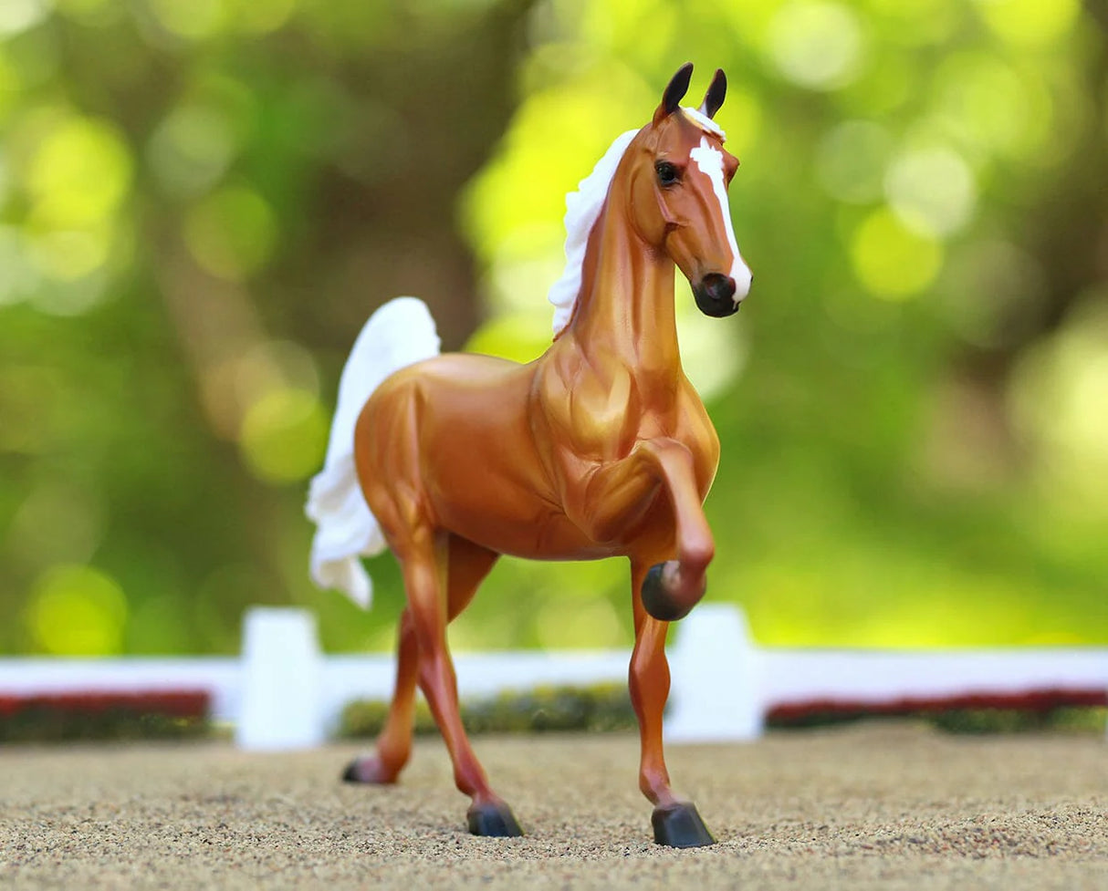 Palomino Saddlebred model horse in mid-step with white mane and tail, set against a blurred green outdoor background.