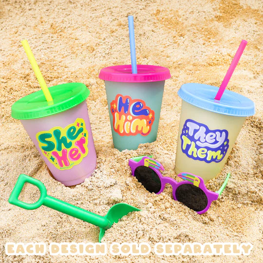 He/Him Pronouns Sticker on colorful cup with straw in sand alongside She/Her and They/Them cups and sunglasses.