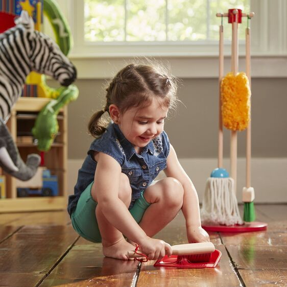 Child playing with Dust Sweep & Mop Set, using hand brush and dust pan with colorful wooden stand in background.