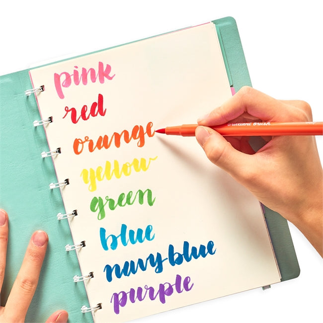 Hand lettering colorful words using Brilliant Brush Markers with soft brush tips on a spiral-bound notebook page.