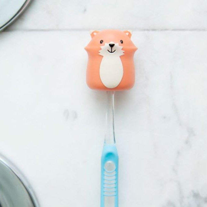 Toothbrush Holder | Fox in orange fox design holding a blue toothbrush on a smooth bathroom surface.