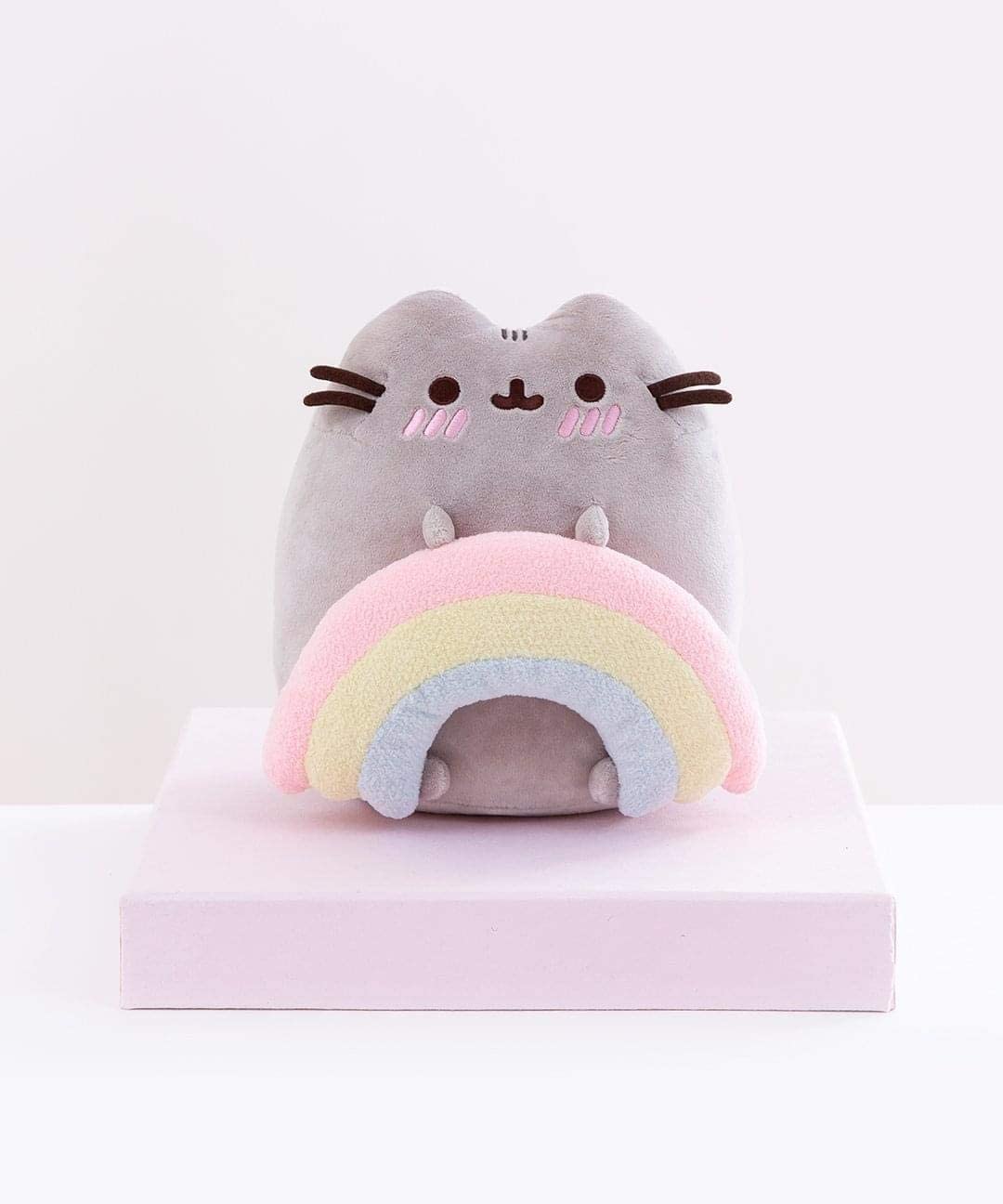 Rainbow Pusheen plush holding a pastel rainbow with embroidered cheeks, standing 9.5 inches tall on a soft pink platform.