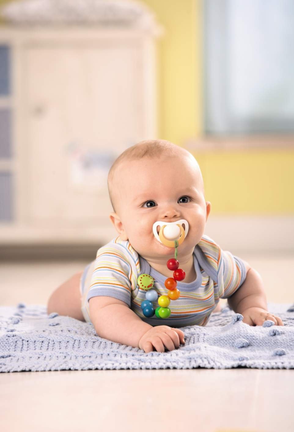 Baby lying on tummy with pacifier attached by colorful Pacifier Chain Rainbow to clothing in bright room