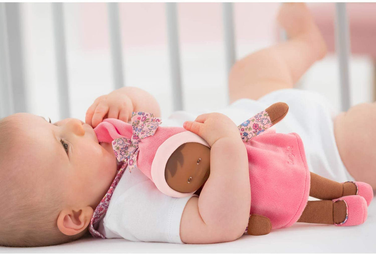 Baby lying in crib holding Miss Floral Sweet Dreams soft doll with stocking cap and floral dress close to face