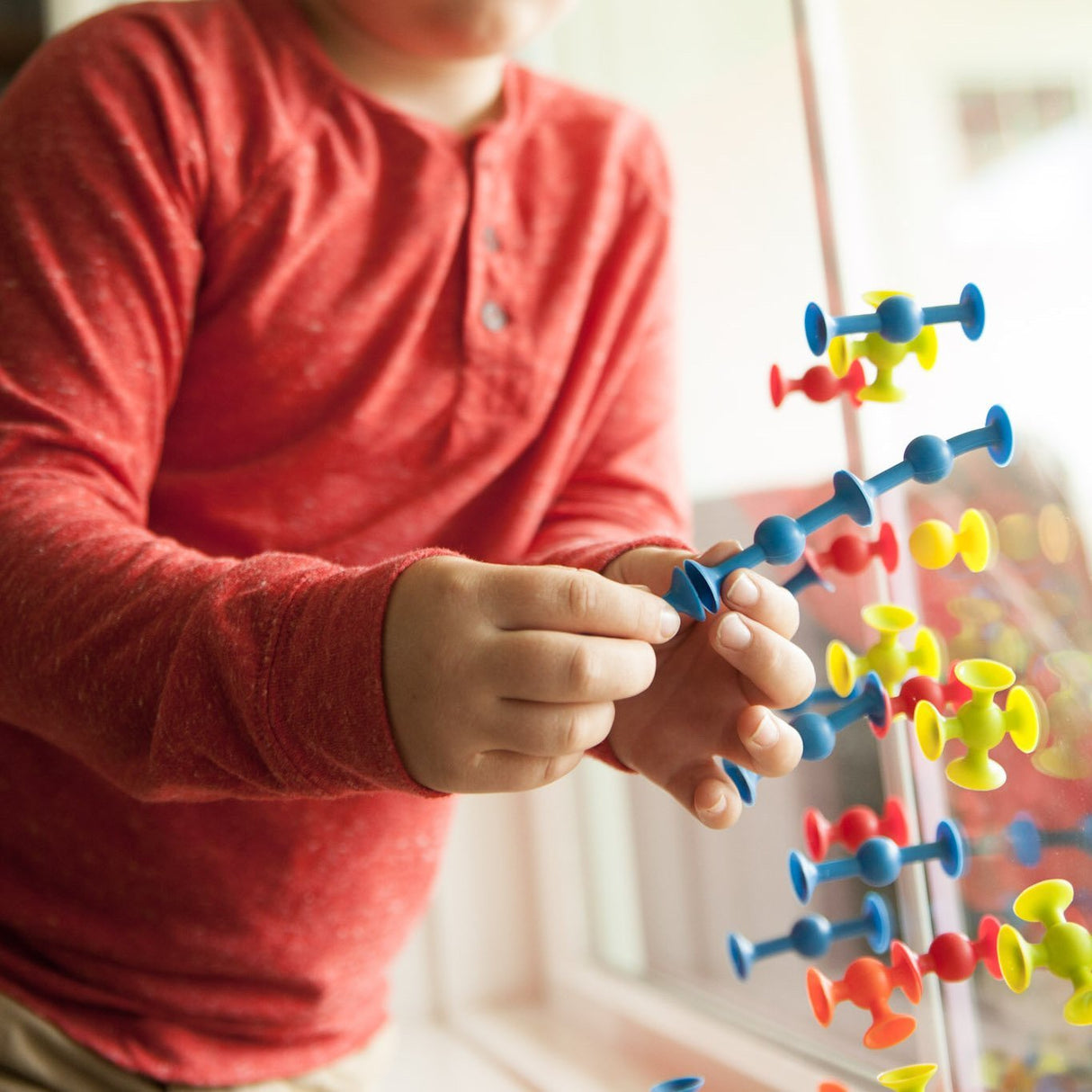 Child playing with colorful Mini Squigz on a window, building creative shapes with the smaller silicone pieces.