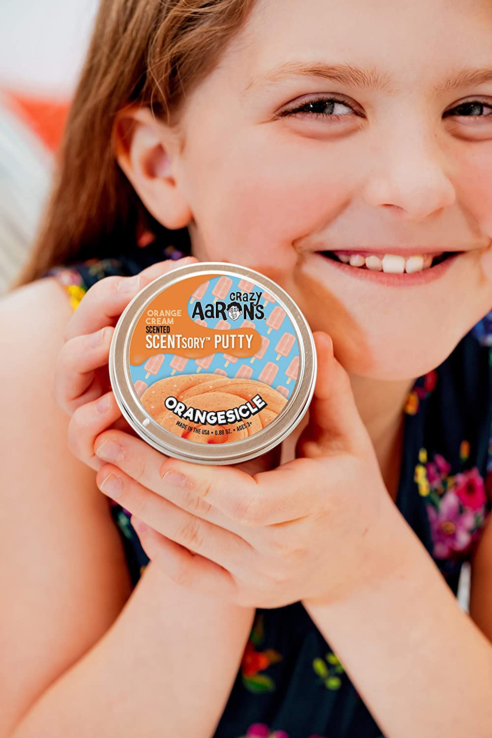 Young girl smiling and holding a tin of SCENTsory Orangesicle Thinking Putty with orange cream scent.