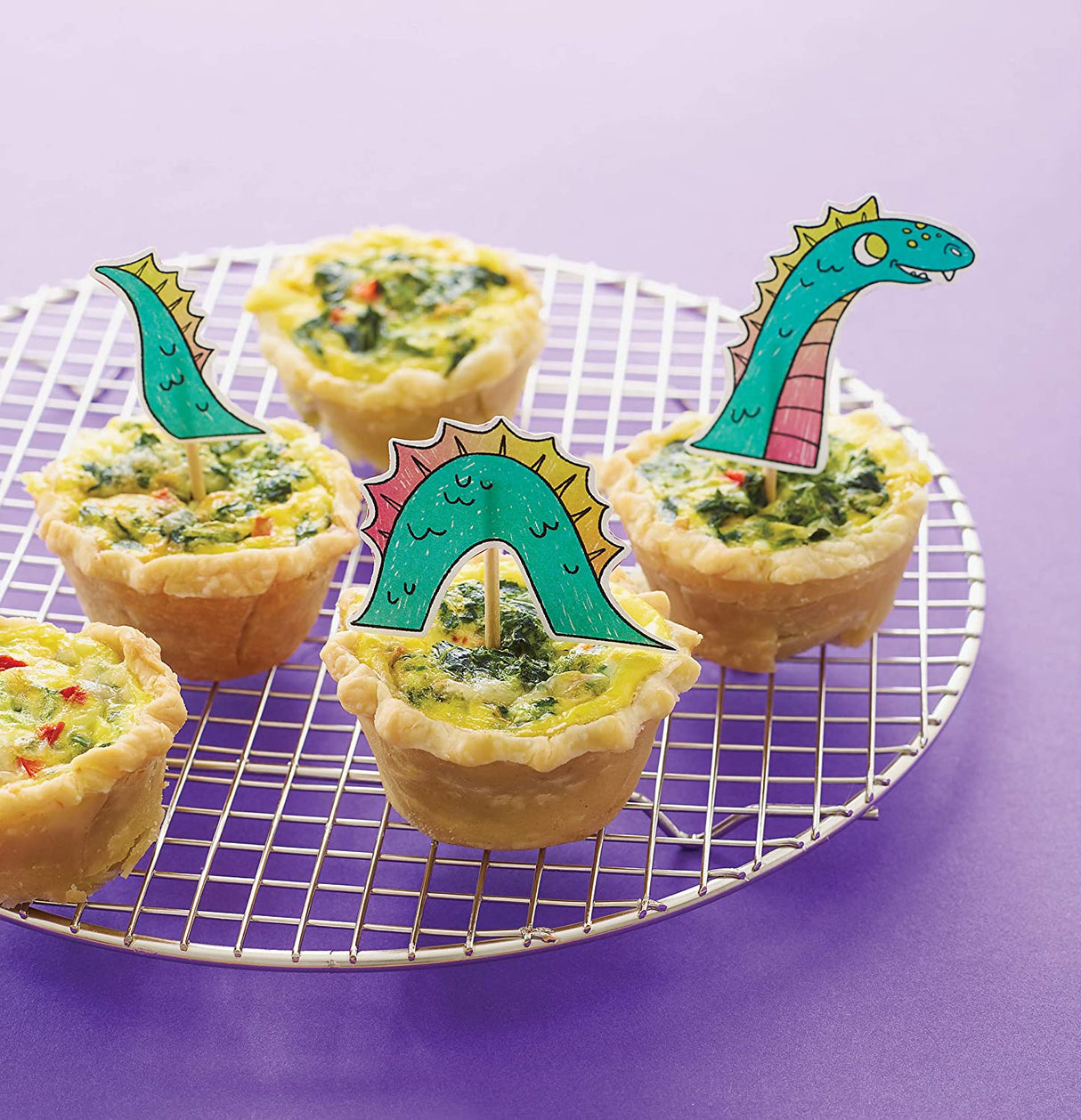 Kids Magical Baking mini pies with colorful dinosaur-themed decorations on cooling rack against purple background