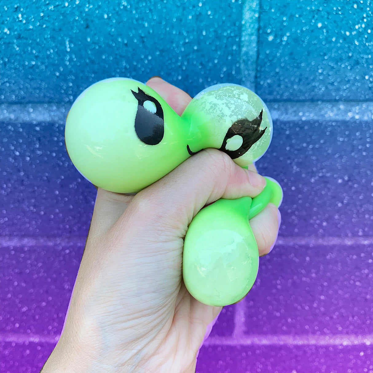 Hand squeezing a green Fun Squad Bubble Blobbie squishy toy with cartoon eyes against a blue and purple wall background